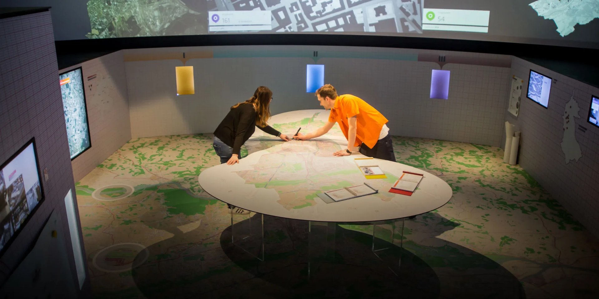 Visitors exploring a large circular map table surrounded by projected spatial data at Ars Electronica