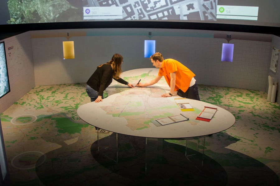 Visitors exploring a large circular map table surrounded by projected spatial data at Ars Electronica