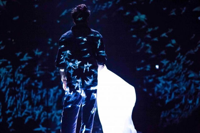 A dancer in Murmur surrounded by blue-white projected fragments on a dark stage
